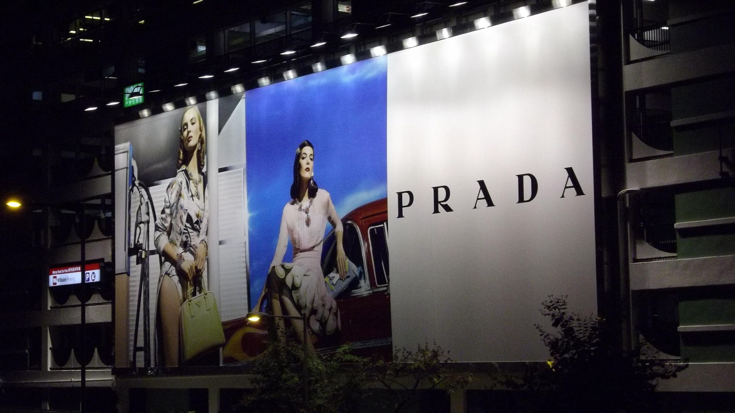 La imagen muestra una gran valla publicitaria de la marca Prada, iluminada por focos en la parte superior. La publicidad presenta dos modelos femeninas con un estilo elegante y sofisticado.  A la izquierda, una modelo viste un conjunto refinado con estampados, sosteniendo un bolso de lujo en tonos claros, con una actitud segura y sofisticada. La imagen tiene una estética vintage y cinematográfica, con una gasolinera de fondo.  En el centro, otra modelo posa con un atuendo en tonos rosados, sentada sobre un coche clásico, con un fondo azul intenso que resalta su presencia. Su expresión y postura transmiten elegancia y exclusividad.  A la derecha, el diseño minimalista de la valla deja un gran espacio en blanco con el nombre “PRADA” en letras negras, destacando la identidad de la marca.  La iluminación nocturna resalta la publicidad, haciéndola aún más impactante y llamativa. La composición refleja la sofisticación y el lujo característicos de la firma Prada.