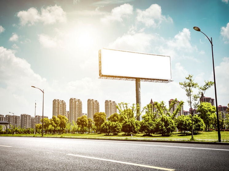 La imagen muestra una carretera amplia y vacía con un gran cartel publicitario en el centro de la escena. El cartel es completamente blanco, sin contenido visible, lo que crea un efecto de vacío o posibilidad de personalización. Al fondo, se observa una ciudad con varios edificios altos, rodeados de áreas verdes con árboles bien cuidados. El cielo es azul con algunas nubes dispersas, y el sol brilla intensamente, creando un efecto de luz que resalta la escena. También hay farolas en los costados de la vía, lo que indica que es una zona urbana moderna y bien iluminada. La imagen transmite una sensación de amplitud y potencial, con el cartel en blanco como punto focal que invita a imaginar qué podría ser anunciado allí